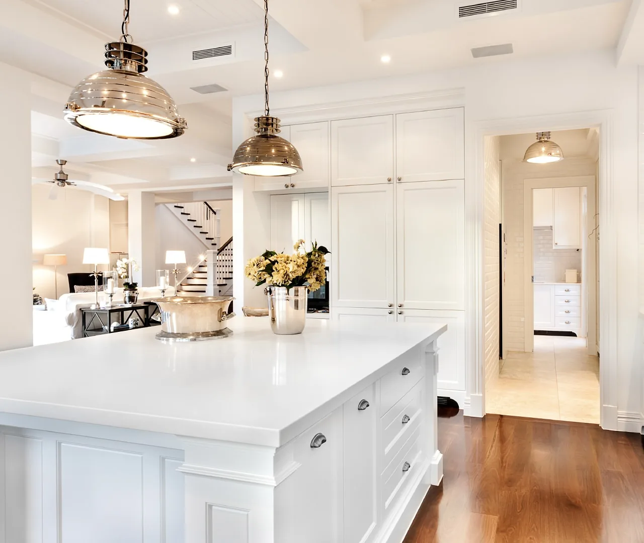 White kitchen with a long island, polished nickel pendants and a stair hall beyond